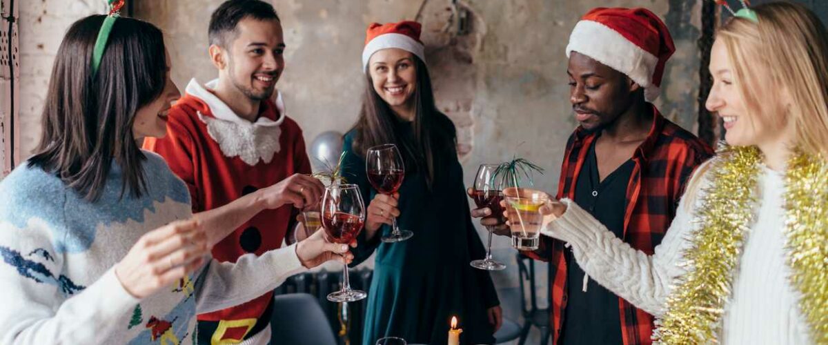 A group of friends wearing holiday sweaters and accessories stand around a table eating snacks, talking, and raising their glasses to cheer