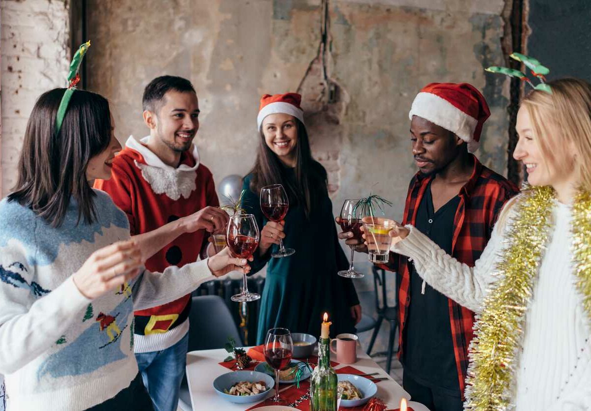 A group of friends wearing holiday sweaters and accessories stand around a table eating snacks, talking, and raising their glasses to cheer