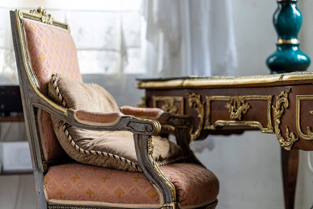 A pink, upholstered chair sitting at an antique desk with gold fixtures. 