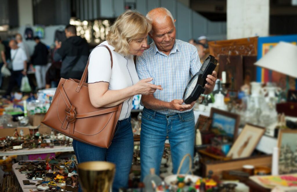 A couple in a flea market examines a clock among a slew of antique items.