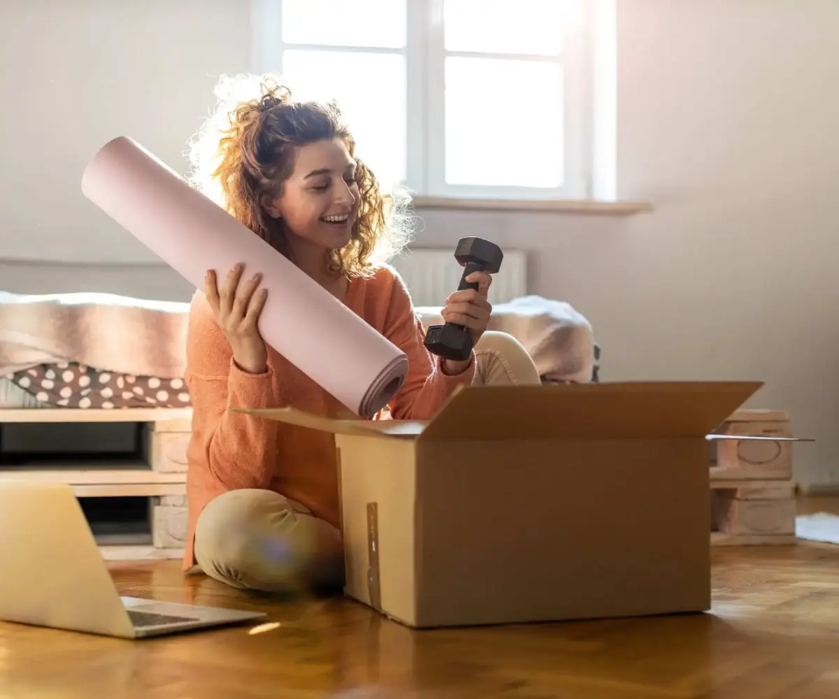 A woman smiles as she opens a package with a yoga mat and free weight.