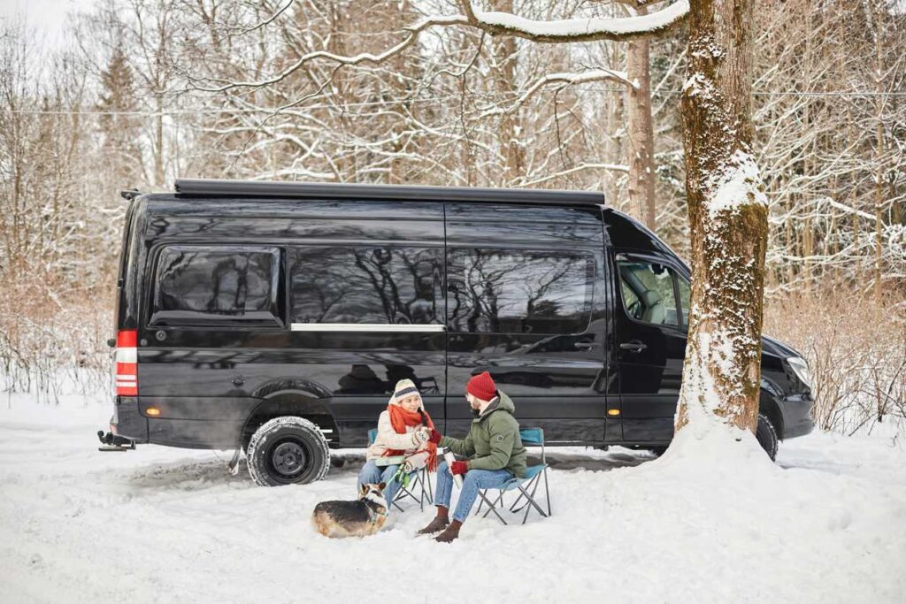 Two people in winter gear and a corgi dog sit in front of a black van in folding chairs in the snow.