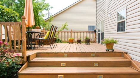 A wooden deck of a home with a table, chairs, outdoor plants, and an umbrella, ready for the summer season.