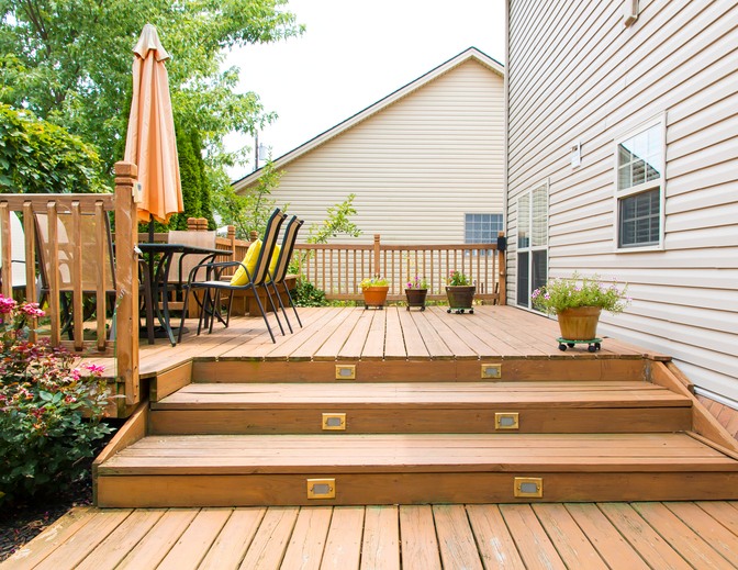 A wooden deck of a home with a table, chairs, outdoor plants, and an umbrella, ready for the summer season.