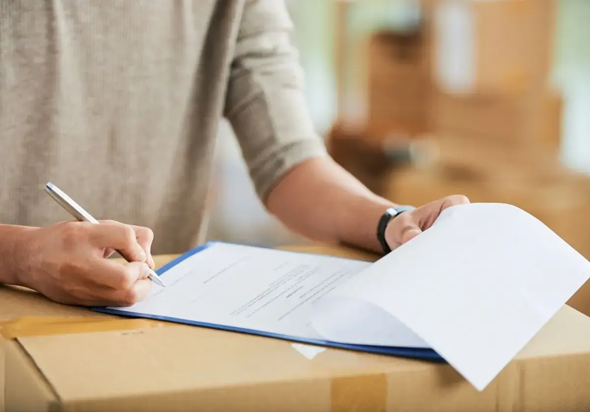 Person signing storage lease agreement on clipboard placed over moving boxes.