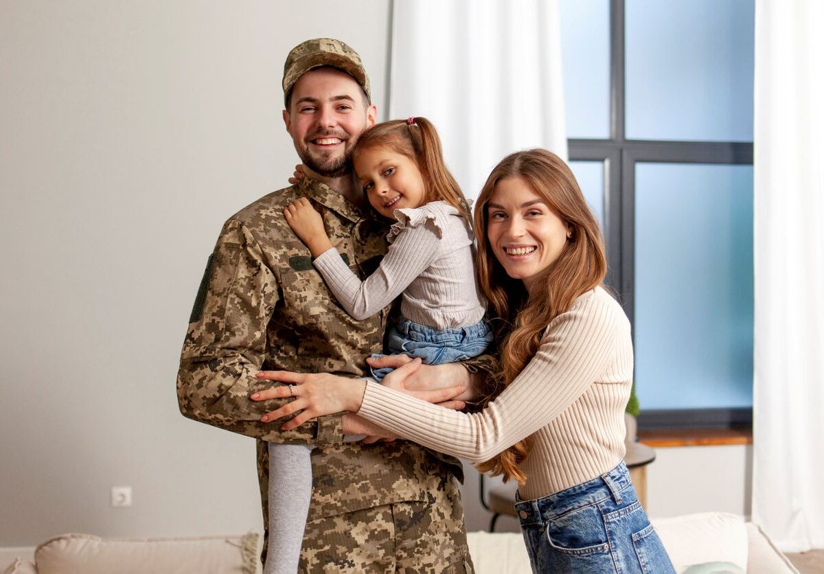 Military man in uniform holding a young girl while a woman hugs them in a living room.