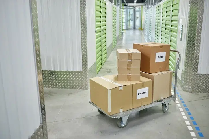 Flatbed cart loaded with labeled cardboard boxes in the hallway of a self storage facility with green unit doors.
