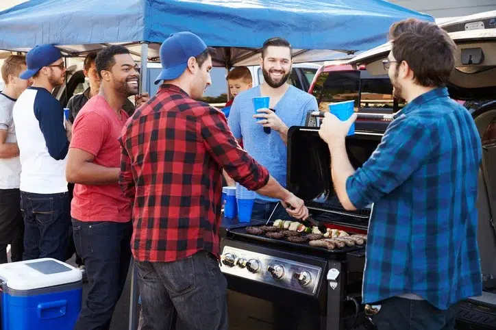 Group of SEC fans grilling burgers and sausages at an outdoor tailgate party under a blue canopy.