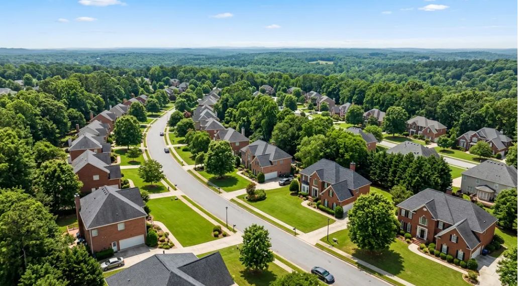 Aerial view of a sunny Georgia suburb with brick homes, tree-lined streets, and green lawns near a wooded horizon.