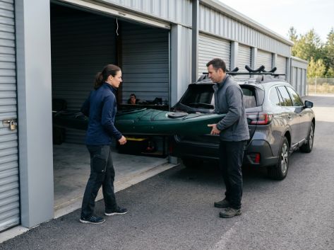 A woman and man unload a kayak from an SUV into an open drive-up storage unit.